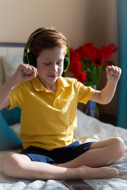 Portrait of a redhaired boy child listens to music in\
headphones sits on the bed and dances contentedly vertical photo\
music lover leisure and entertainment