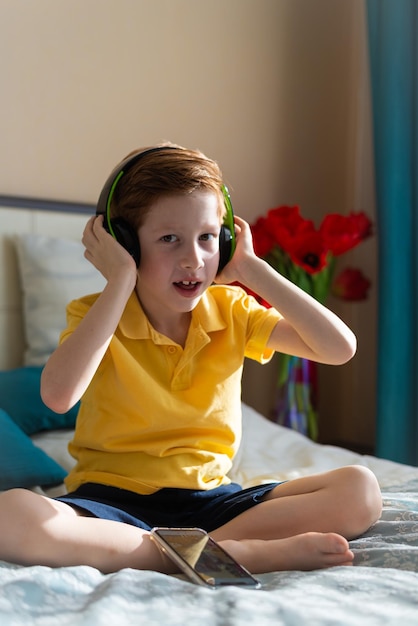 Portrait of a redhaired boy child listens to music in\
headphones sits on the bed dances vertical photo music lover\
leisure and entertainment