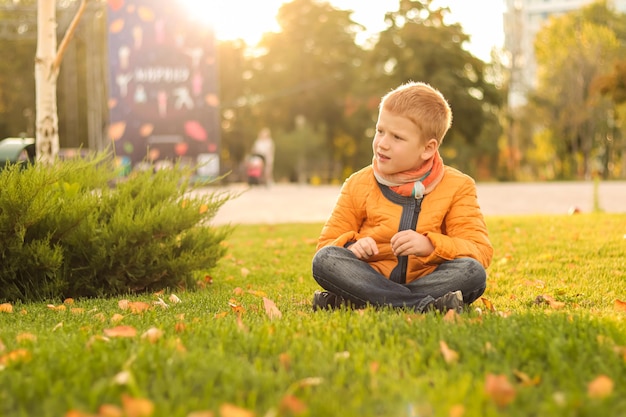 Portrait of a redhaired teenager boy in yellow jacket sitting on the grass in city park at sunset