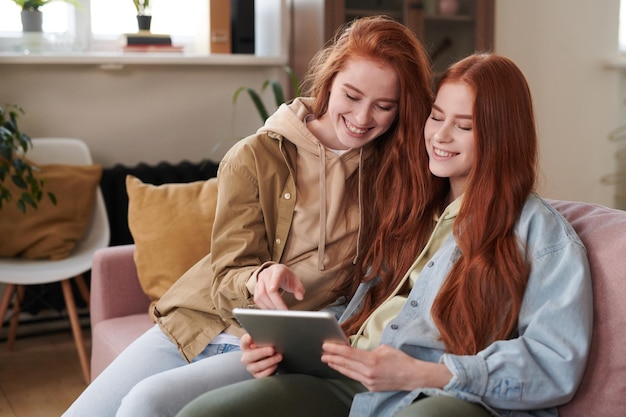 Portrait of redhaired twin sisters spending free time together sitting on sofa in living room enjoying surfing Internet using digital tablet