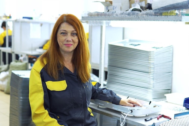 Ritratto di lavoratore di fabbrica donna dai capelli rossi seduto vicino alla produzione catena di montaggio presso lo stabilimento per la produzione di prodotti elettrici