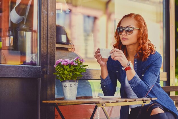 Portrait of redhead female in sunglasses, drinks coffee in a cafe on a street.