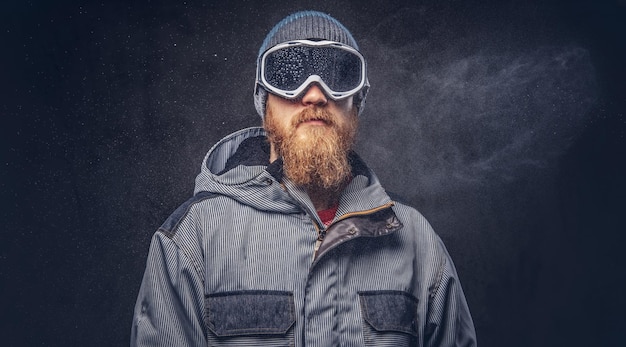 Портрет рыжего сноубордиста с окладистой бородой в зимней шапке и защитных очках, одетого в сноубордическую куртку, позирующего в студии и отводящего взгляд. Изолированные на сером фоне.