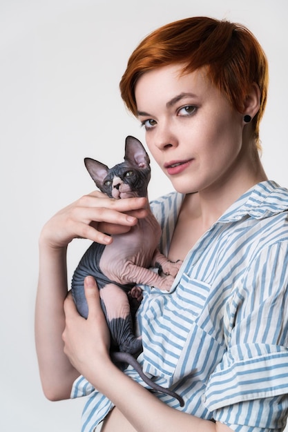 Portrait of redhead young woman holding Sphynx Cat in hands and looking at camera