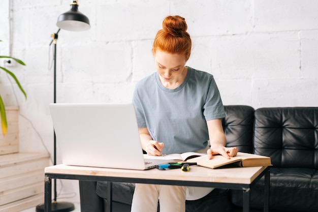 Portrait of redhead young woman student noting into the workbook important information from book