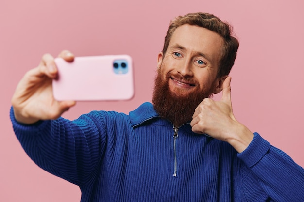 Foto ritratto di un uomo dai capelli rossi con il telefono in mano che scatta selfie e foto sul suo telefono con un sorriso su un blogger di sfondo rosa