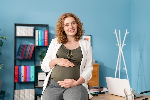 Портрет рыжеволосой беременной деловой женщины, сидящей за деревянным столом в офисе беременной женщины