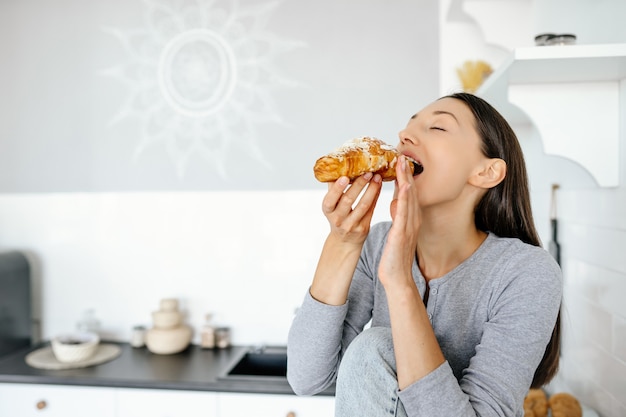 Portrait of rejoicing woman eats tasty croissant at home. Unhealthy food concept.