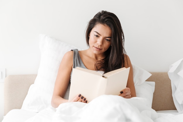 Portrait of relaxed adorable woman with long brown hair resting in comfortable bed after sleep, and reading book