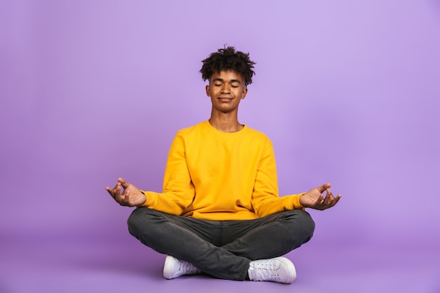 Portrait of relaxed african american boy sitting in lotus pose and meditating with closed eyes, isolated over violet background