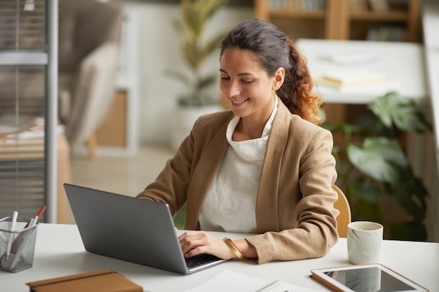 Portrait of relaxed elegant businesswoman smiling happily and using laptop while enjoying work in office, copy space