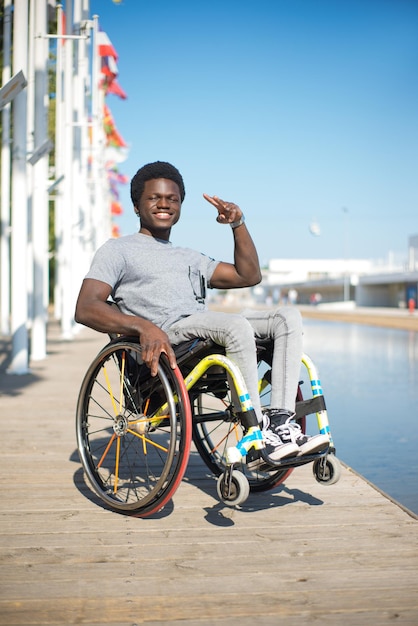 Portrait of relaxed man in wheelchair. African American man in casual clothes on embankment, showing victory sign. Blue sky and flags in background. Portrait, beauty, happiness concept