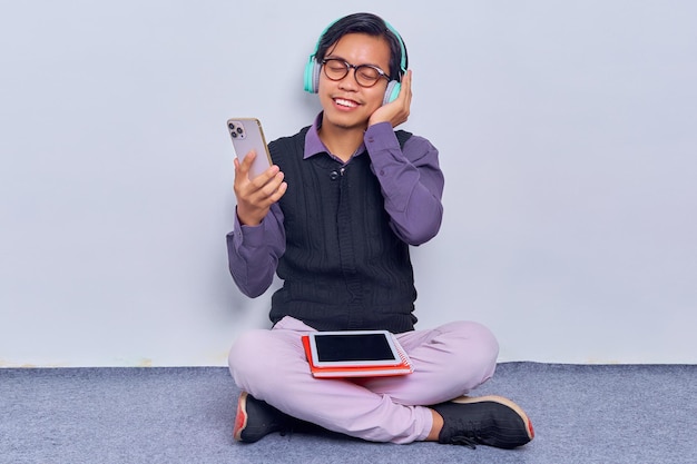 足を組んで白い背景で隔離の床に座って携帯電話を保持している音楽のヘッドフォンを聞いてメガネでリラックスしたプロの若いアジア人の肖像画