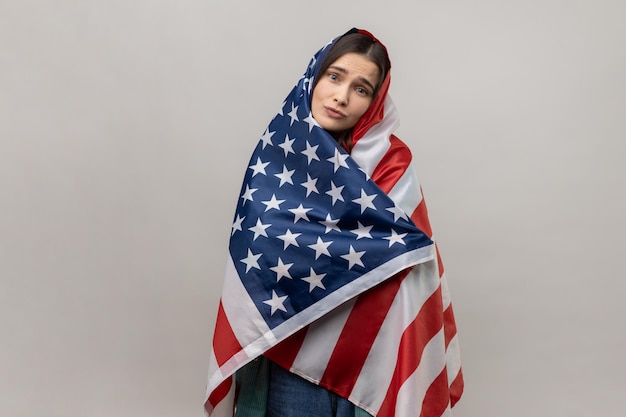 Portrait of relaxed young adult woman standing wrapped in USA flag looking at camera with pout lips