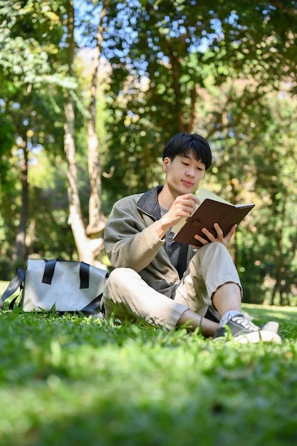 Portrait Relaxed young Asian male college student chilling in the greenery park reading a book