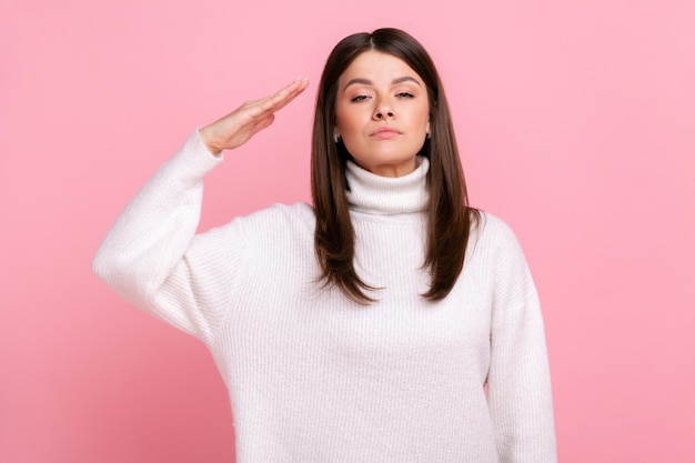 Portrait of responsible serious woman saluting commander listening order with obedient expression wearing white casual style sweater Indoor studio shot isolated on pink background