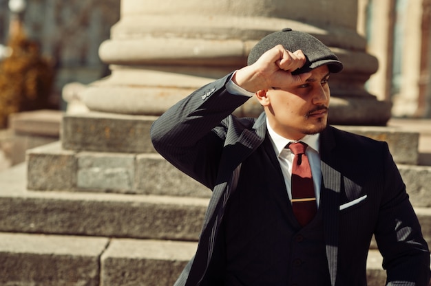 Portrait of retro 1920s english arabian business man wearing\
dark suit, tie and flat cap near old columns.