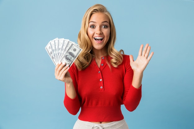 Portrait of rich blond woman 20s wearing red shirt holding fan of dollar money, isolated over blue wall