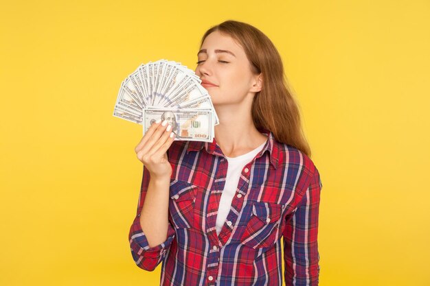 Photo portrait of rich greedy woman in casual checkered shirt holding money smelling dollar banknotes with pleased expression enjoying wealthy life indoor studio shot isolated on yellow background