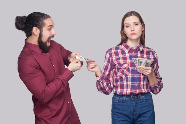 Portrait of rich serious woman with fan of money, sharing with amazed or surprised hungry man. standing and looking at camera like a boss. indoor studio shot, isolated on grey background.