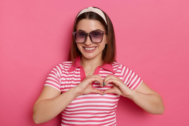 Портрет романтичной красивой женщины в полосатой футболке и солнцезащитных очках, нежно смотрящей в камеру и показывающей жест любви, позирующей изолированно на розовом фоне