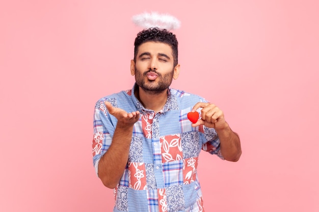 Portrait of romantic guy with beard with closed eyes sending air kissing to you, holding small red heart in hands, expressing love and romance. Indoor studio shot isolated on pink background.