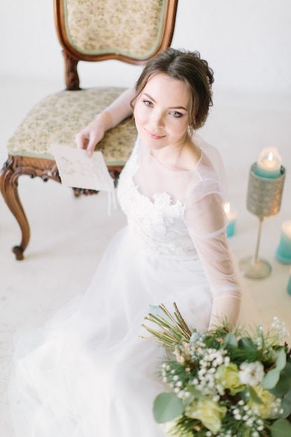 Портрет романтичной нежной невесты в белом длинном свадебном платье, сидящей рядом со стулом и свечами на полу и держащей букет цветов