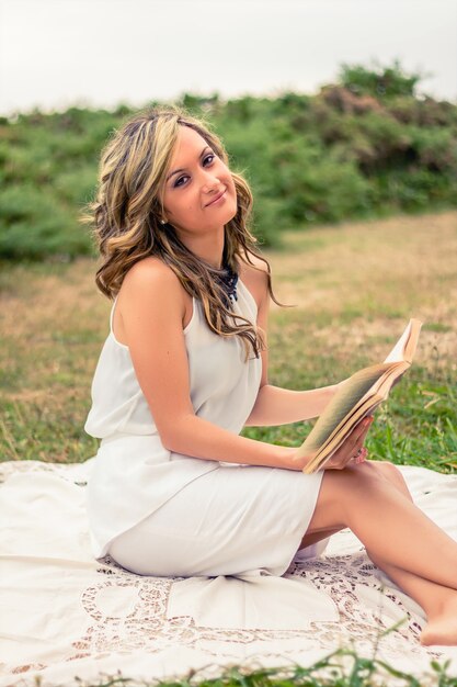 Portrait of romantic young woman reading a book sitting over the grass. Relax outdoor time concept.