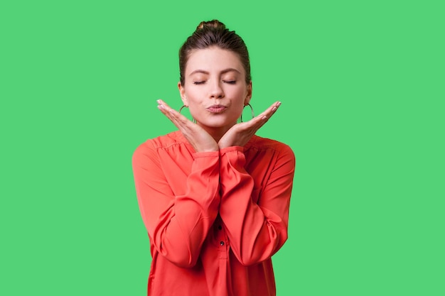 Portrait of romantic young woman with bun hairstyle big earrings and in red blouse standing with closed eyes blowing air kiss with pout lips flirt indoor studio shot isolated on green background