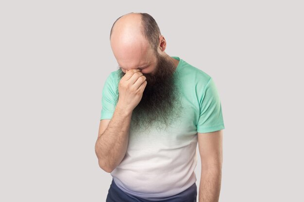 Portrait of sad alone depressed middle aged bald man with long beard in light green tshirt standing holding his head down and crying indoor studio shot isolated on grey background