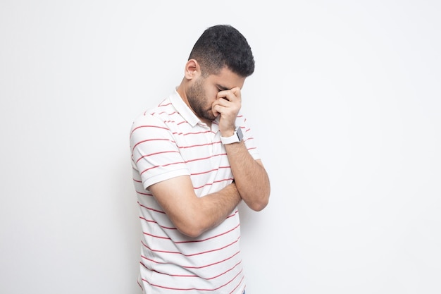 Portrait of sad alone handsome bearded young man in striped t-shirt standing, holding his head down and crying. indoor studio shot, isolated on white background.