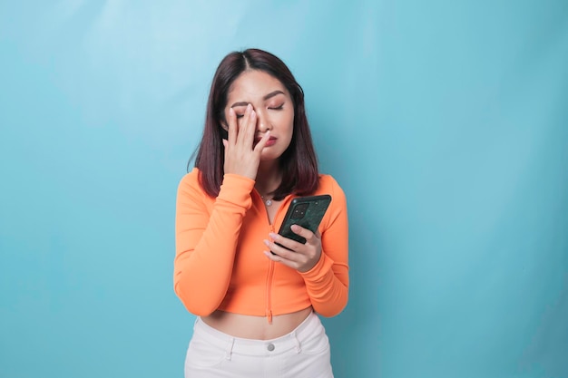 A portrait of a sad Asian woman looking at her smartphone over blue background