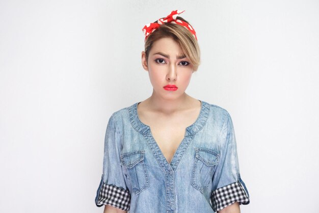 Portrait of sad beautiful young woman in casual blue denim shirt with makeup and red headband standing and looking at camera with unhappy face. indoor studio shot, isolated on white background.