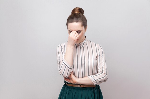 Photo portrait of sad depressed young woman in striped shirt and green skirt with collected ban hairstyle, standing, holding head hown and crying. indoor studio shot, isolated on grey background.