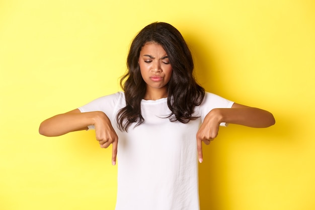 Portrait of sad and disappointed african-american girl, complaining about something bad, looking and pointing fingers down with displeased face, standing over yellow background