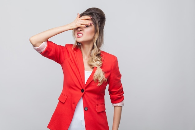 Portrait of sad lose beautiful business lady with hairstyle and makeup in red fancy blazer, standing holding hand on her forehead. indoor studio shot, isolated on grey background.