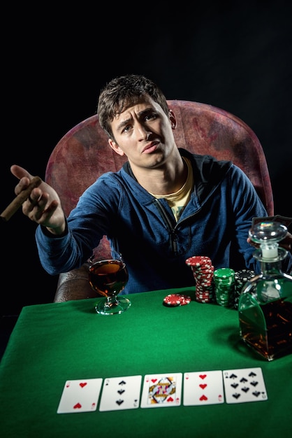 portrait of sad man sitting at poker table with whiskey and cigar against black background
