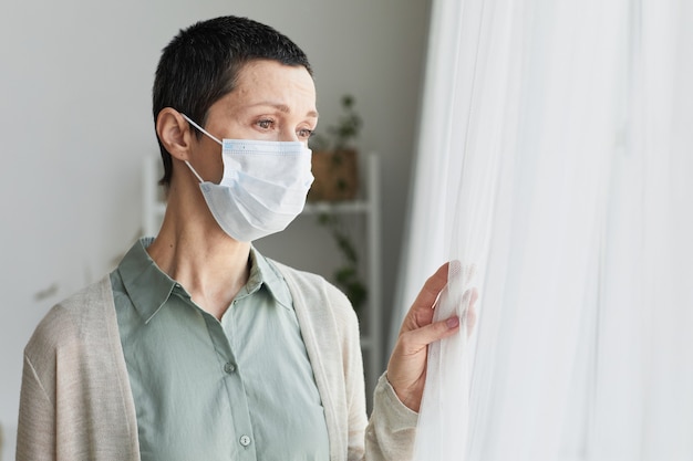 Portrait of sad mature woman wearing mask at home and looking at window, covid, social distancing concept