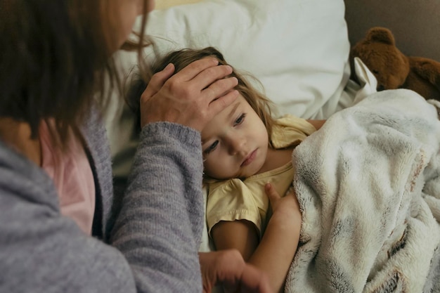 Portrait of sad sick little girl and her mother touching daughter's forehead