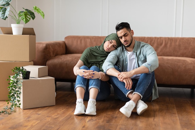 Portrait of sad tired arab family of two people sitting on the floor, leaning on the couch in living room with cardboard boxes around. Exhausted man and woman in headscarf packing or unpacking things