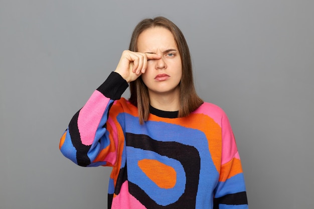 Portrait of sad tired sick woman wearing colorful sweater rubbing her eye looking at camera frowning face being overworked posing isolated over gray background