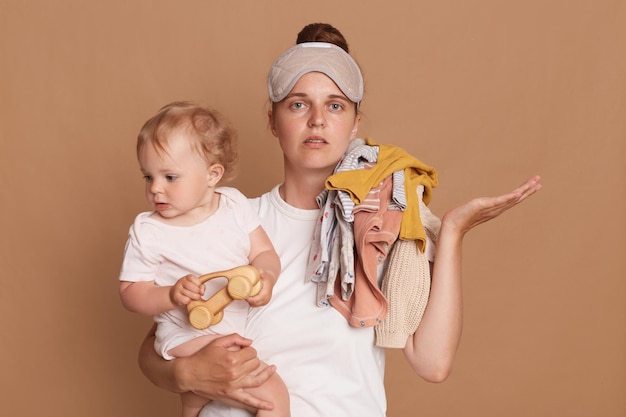 Ritratto di triste sconvolto stressato giovane madre con acconciatura da panino che tiene la figlia del bambino e vestiti sulla spalla spalancando il palmo da parte in posa isolata su sfondo marrone