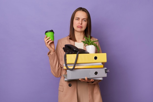 Portrait of sad worried tired exhausted caucasian woman wearing official style jacket holding folders isolated over purple background drinking takeaway coffee needs energy