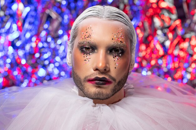 Portrait of sad young bearded man with glitter makeup wearing white fluffy ruff against tinsel background, New Year concept