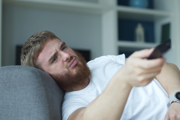 Портрет грустного молодого человека в повседневной одежде, сидящего и лежащего на диване в гостиной и смотрящего телевизор