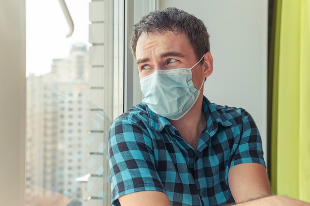 Portrait of a sad young man in a medical mask who looks out the window through the window, he observes quarantine. lockdown