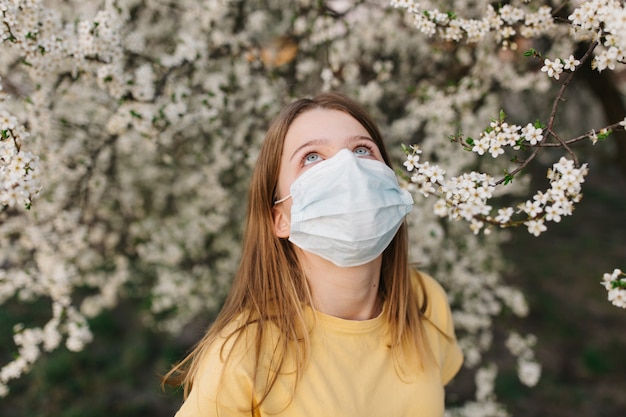 Портрет грустно молодая женщина в защитной медицинской маске с цветами возле цветущего дерева в весеннее время. Концепция весенней аллергии
