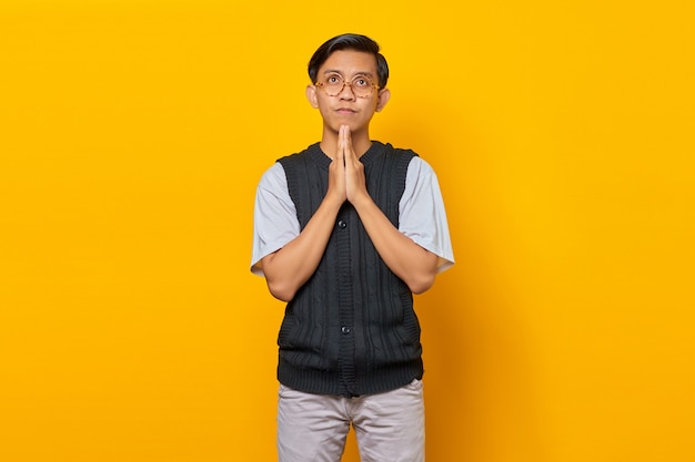 Portrait of sadness young asian man showing praying gesture on yellow background