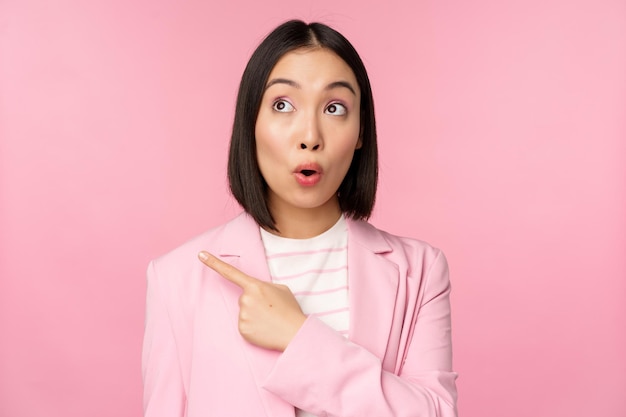 Portrait of saleswoman korean businesswoman pointing and looking left with surprised intrigued face expression posing in suit over pink background