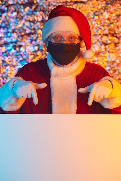 Foto ritratto di babbo natale in maschera protettiva e che punta al grande cartello vuoto